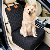 A small fluffy tan dog sits on a black quilted car seat cover installed in a beige car interior.